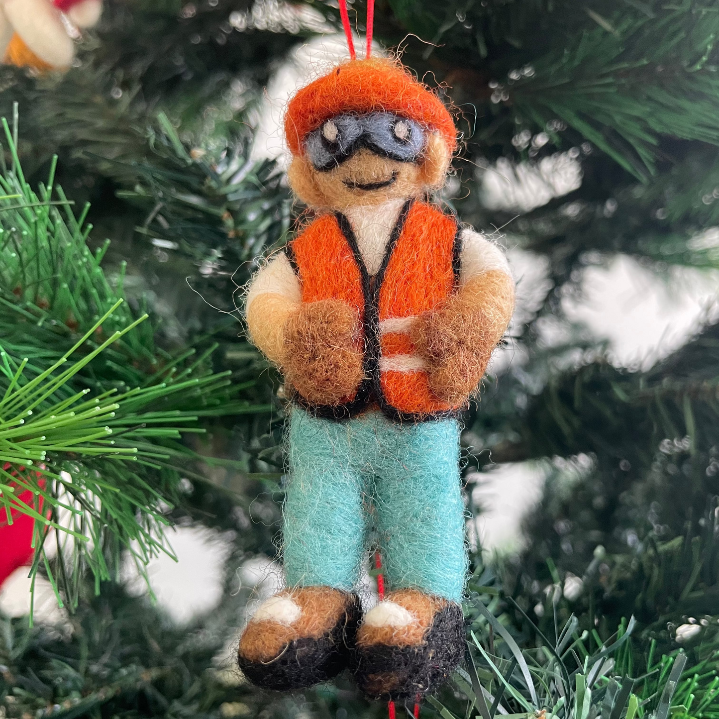 A handcrafted felt Christmas ornament shaped like a construction worker hanging on a green Christmas tree. The ornament features an orange hat and vest, light blue pants, and brown shoes, with a red hanging string at the top.