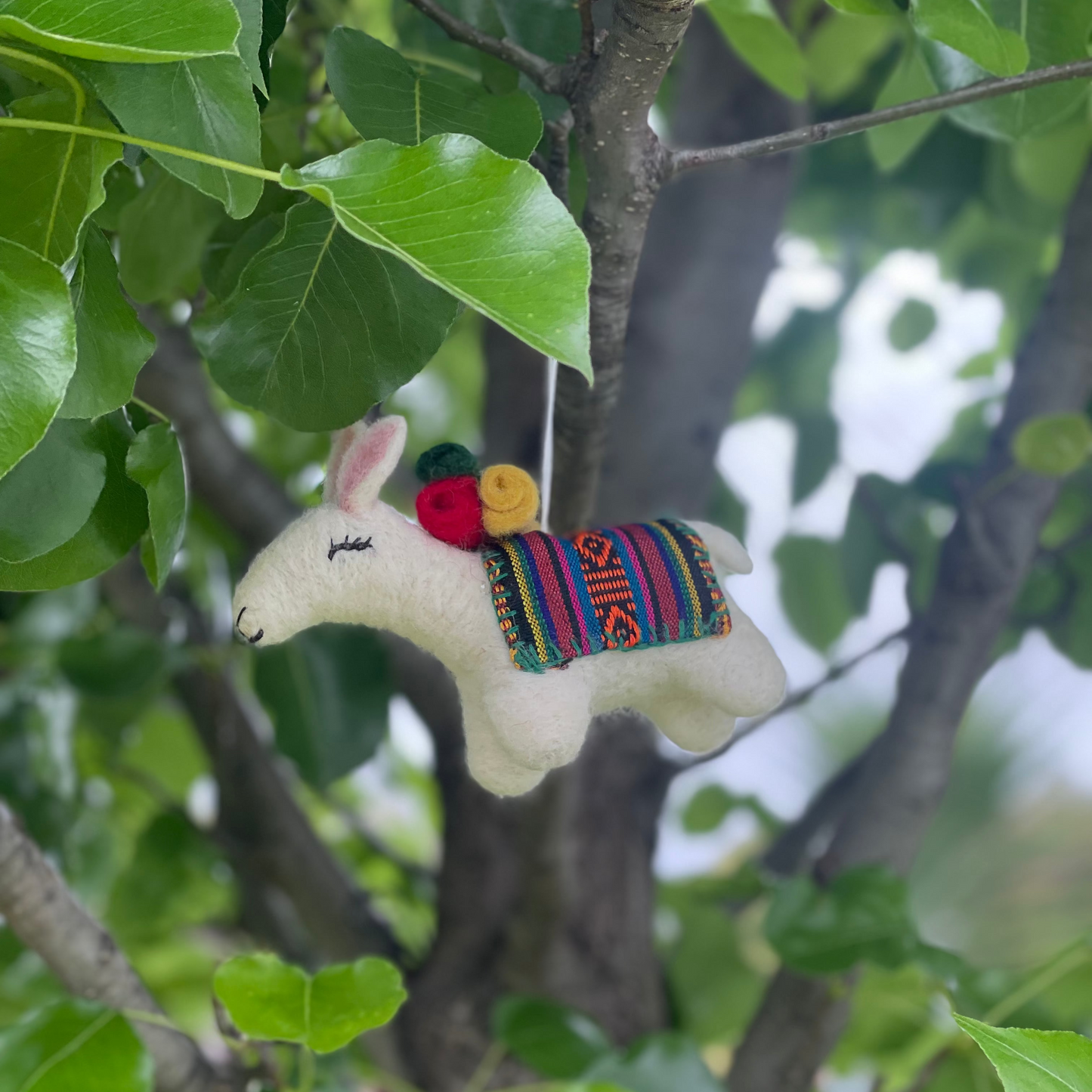 A small white llama or alpaca-shaped hanging ornament with a colorful striped saddle and red, yellow, and green accents, photographed hanging from a tree branch with green leaves.